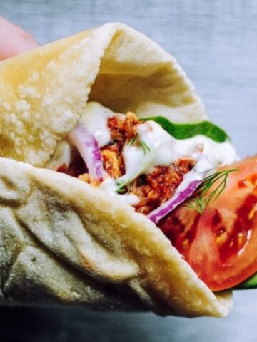 A hand holding a pita wrap filled with seasoned meat, tomato, onion, lettuce, and sauce on a gray surface.