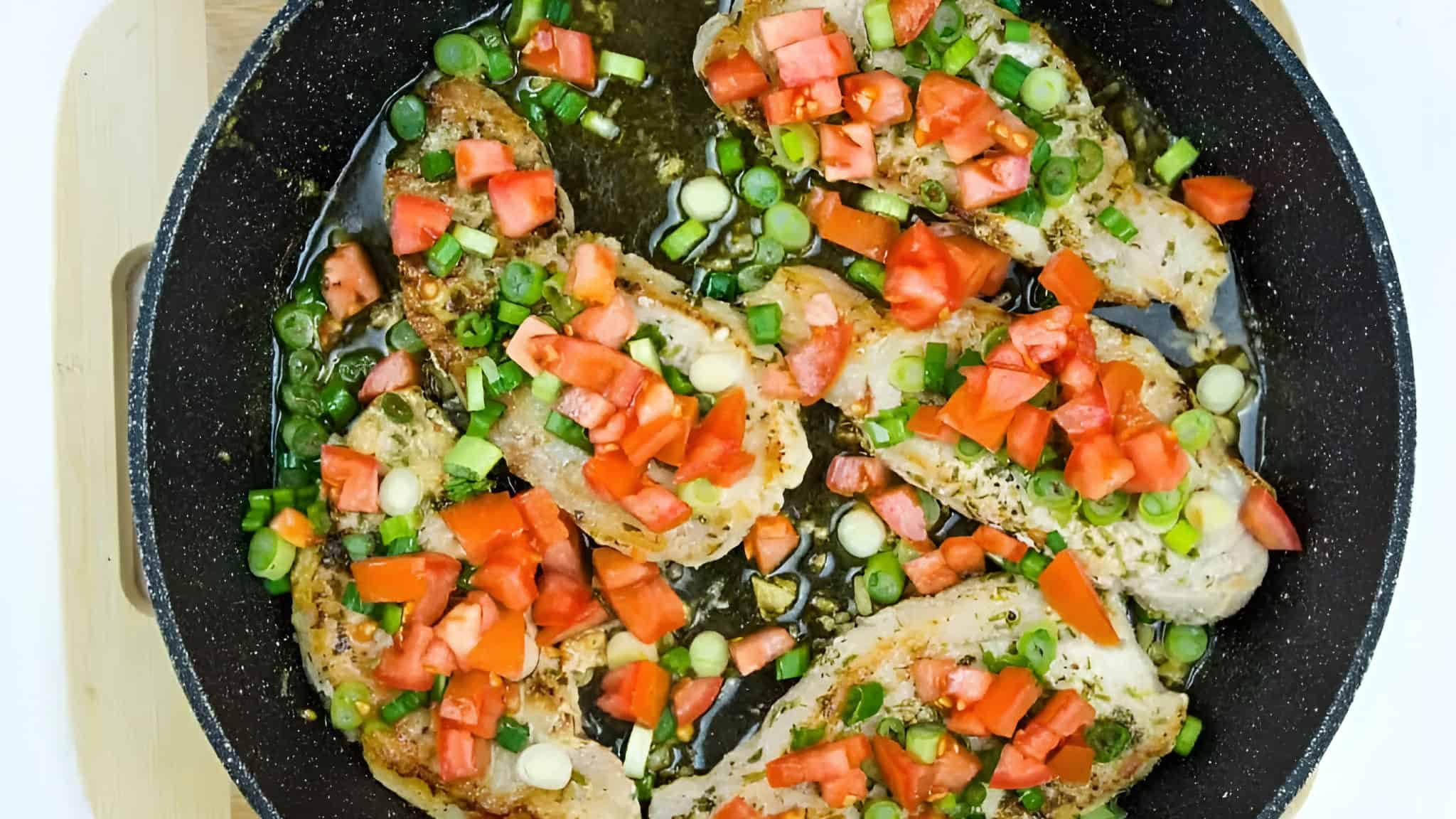 Chicken breasts cooking in a pan, topped with chopped tomatoes and green onions.