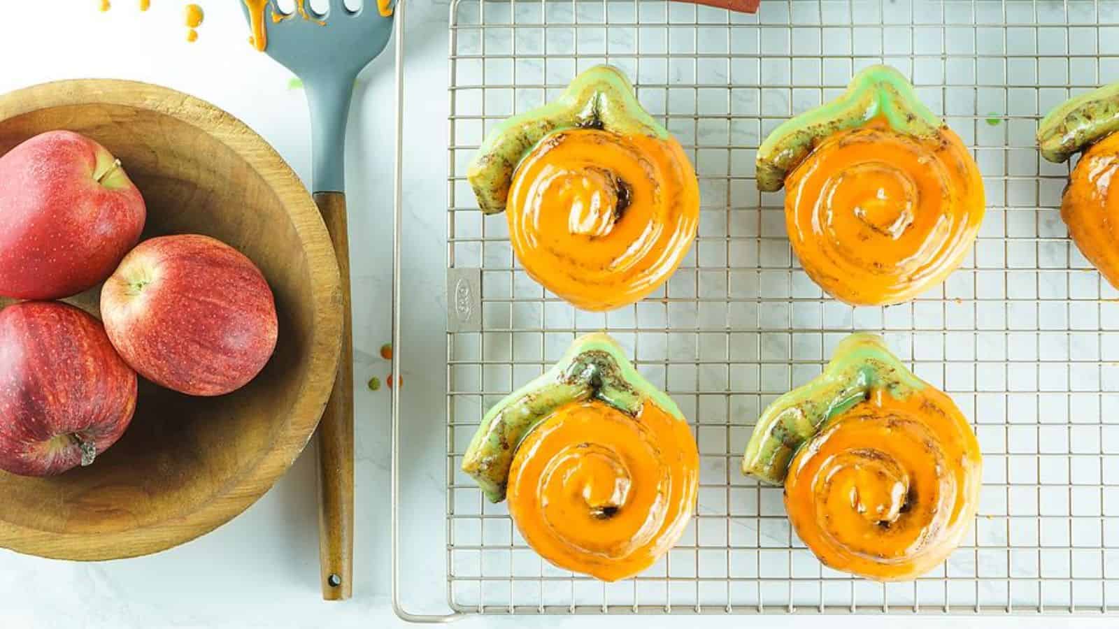 Six glazed cinnamon rolls with green stems on a cooling rack, resembling pumpkins. A bowl of red apples and a spatula are nearby.