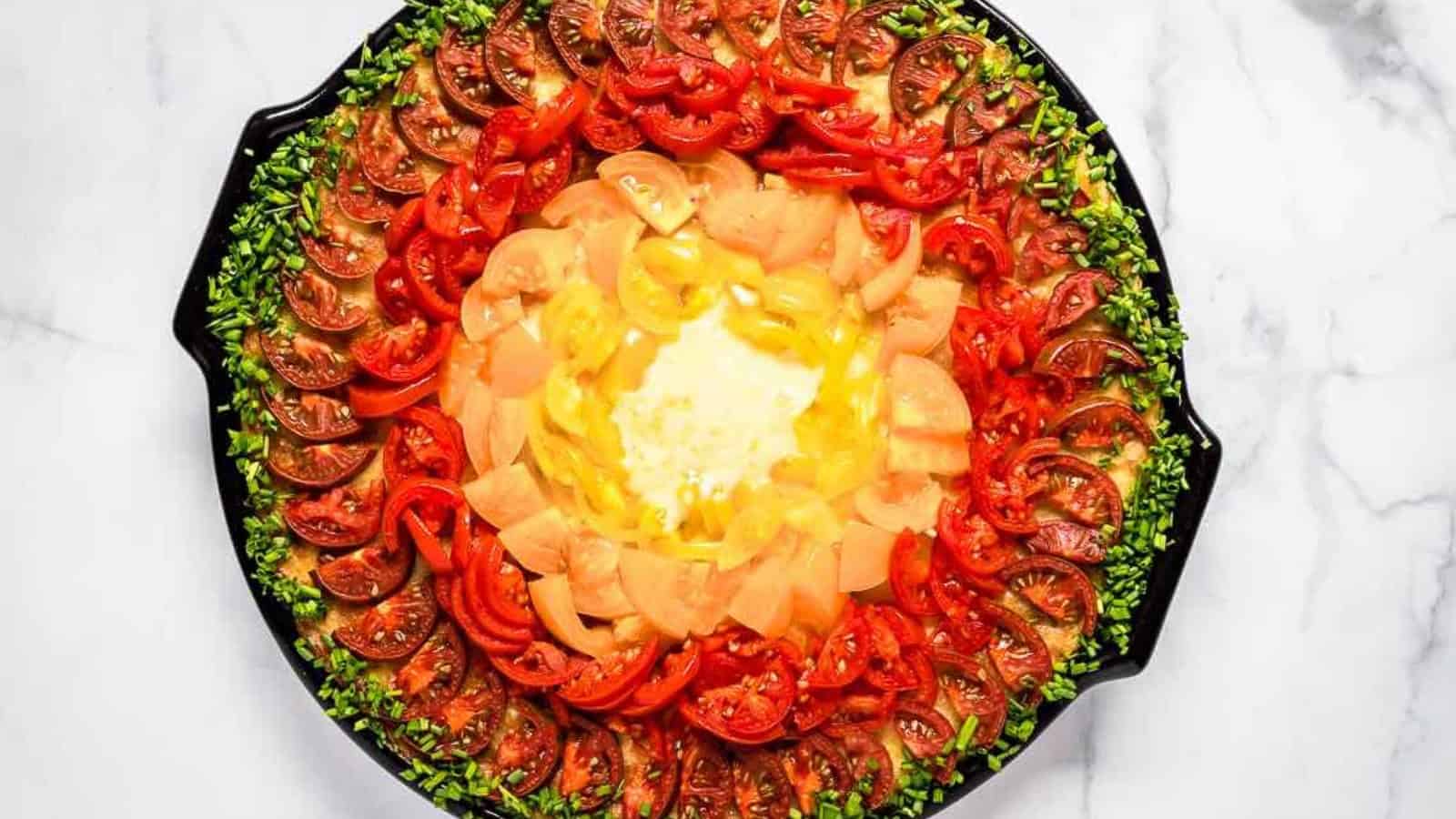 Colorful vegetable arrangement on a plate with concentric circles of chopped greens, sliced red tomatoes, yellow and orange bell peppers on a white marble surface.