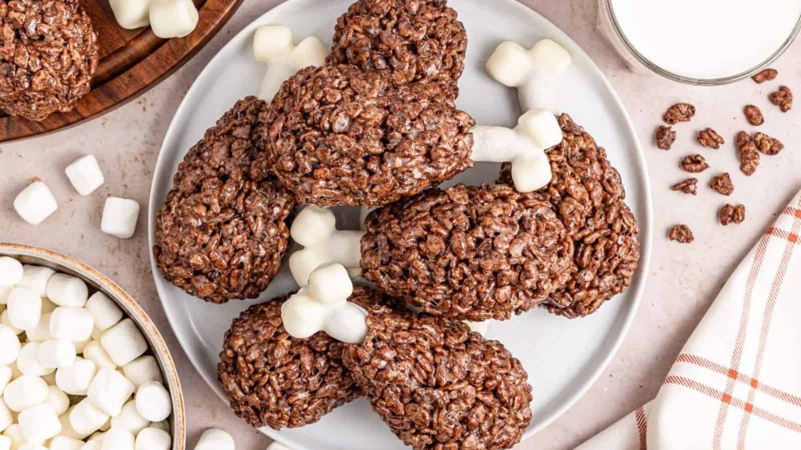 Crispy rice treats shaped like turkey legs with marshmallow "bones" on a white plate, surrounded by mini marshmallows and glass of milk.