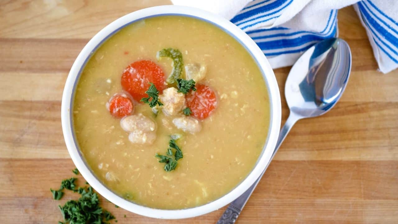 A bowl of soup with carrots, beans, and garnished herbs on a wooden table. A spoon and a striped cloth are beside it.