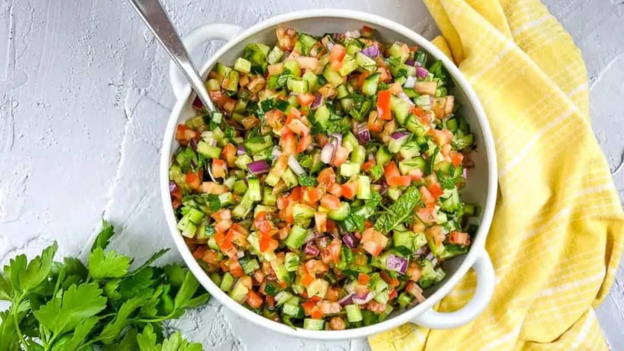 A bowl of diced cucumber, tomato, red onion, and herbs with a spoon, next to a yellow cloth and parsley.