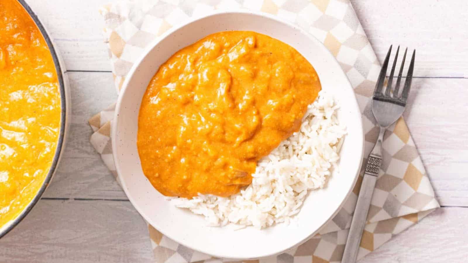 A bowl of creamy orange curry served over white rice, placed on a patterned napkin with a fork beside it.