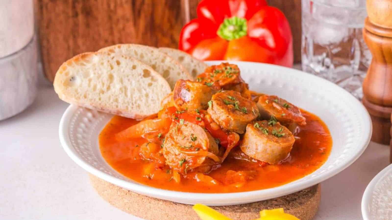 Plate of sliced sausage in tomato sauce with herbs, served with two slices of bread. A red bell pepper and glass of water are in the background.