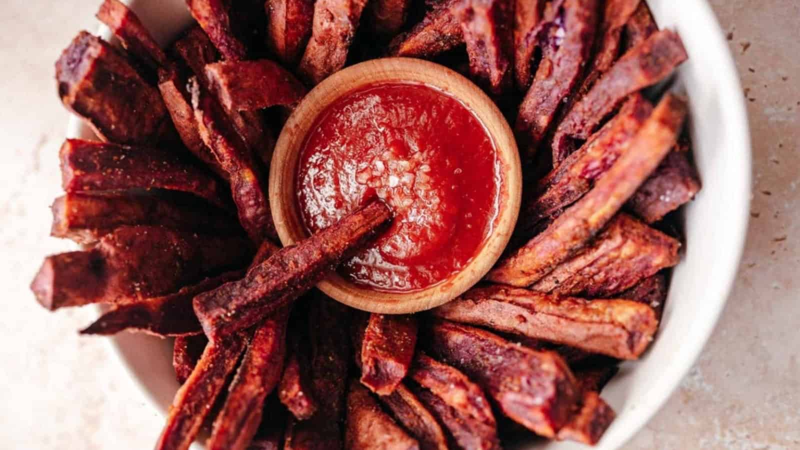 A bowl of crispy sweet potato fries arranged around a small wooden bowl of red dipping sauce.