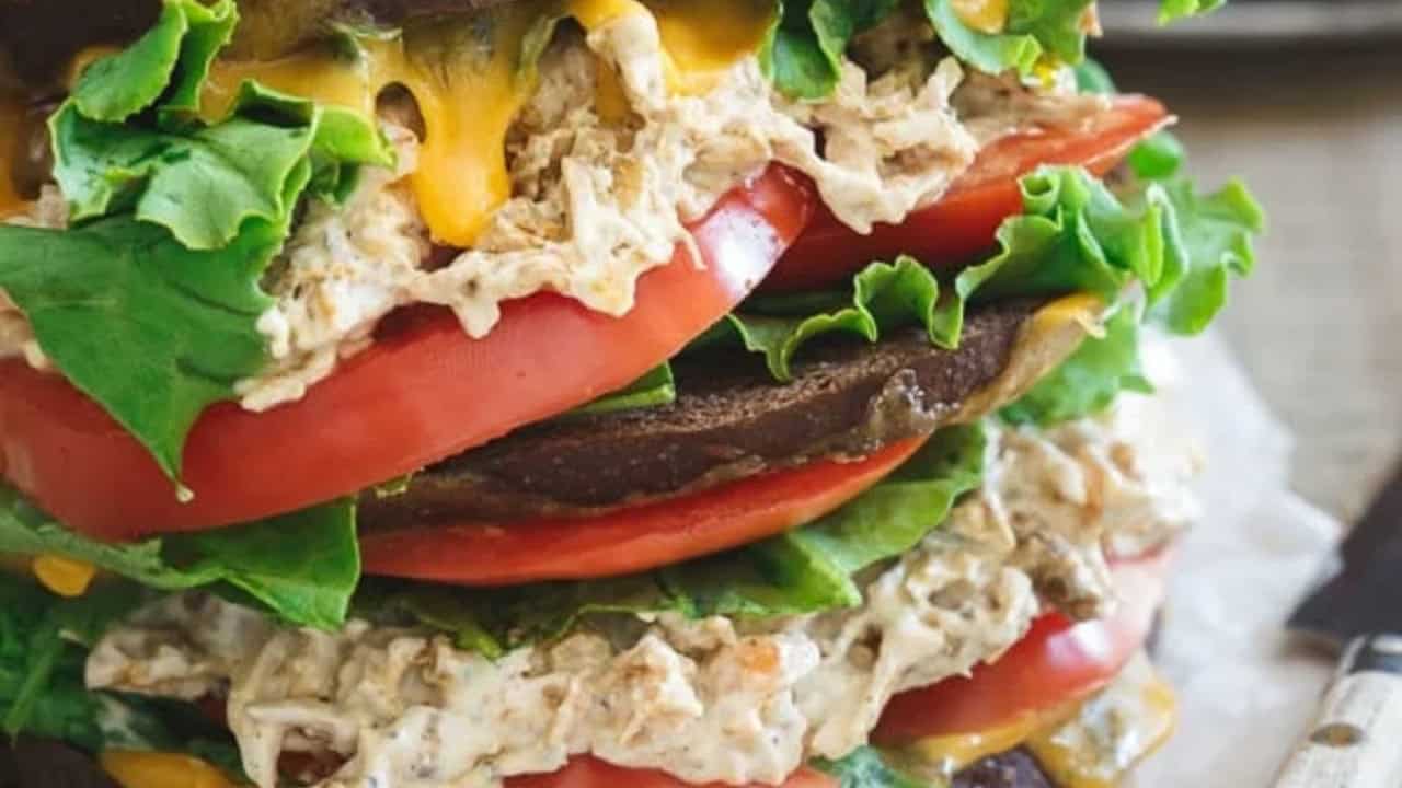A close-up of a sandwich stacked with portobello mushrooms, lettuce, tomato slices, shredded chicken, and melted cheese.