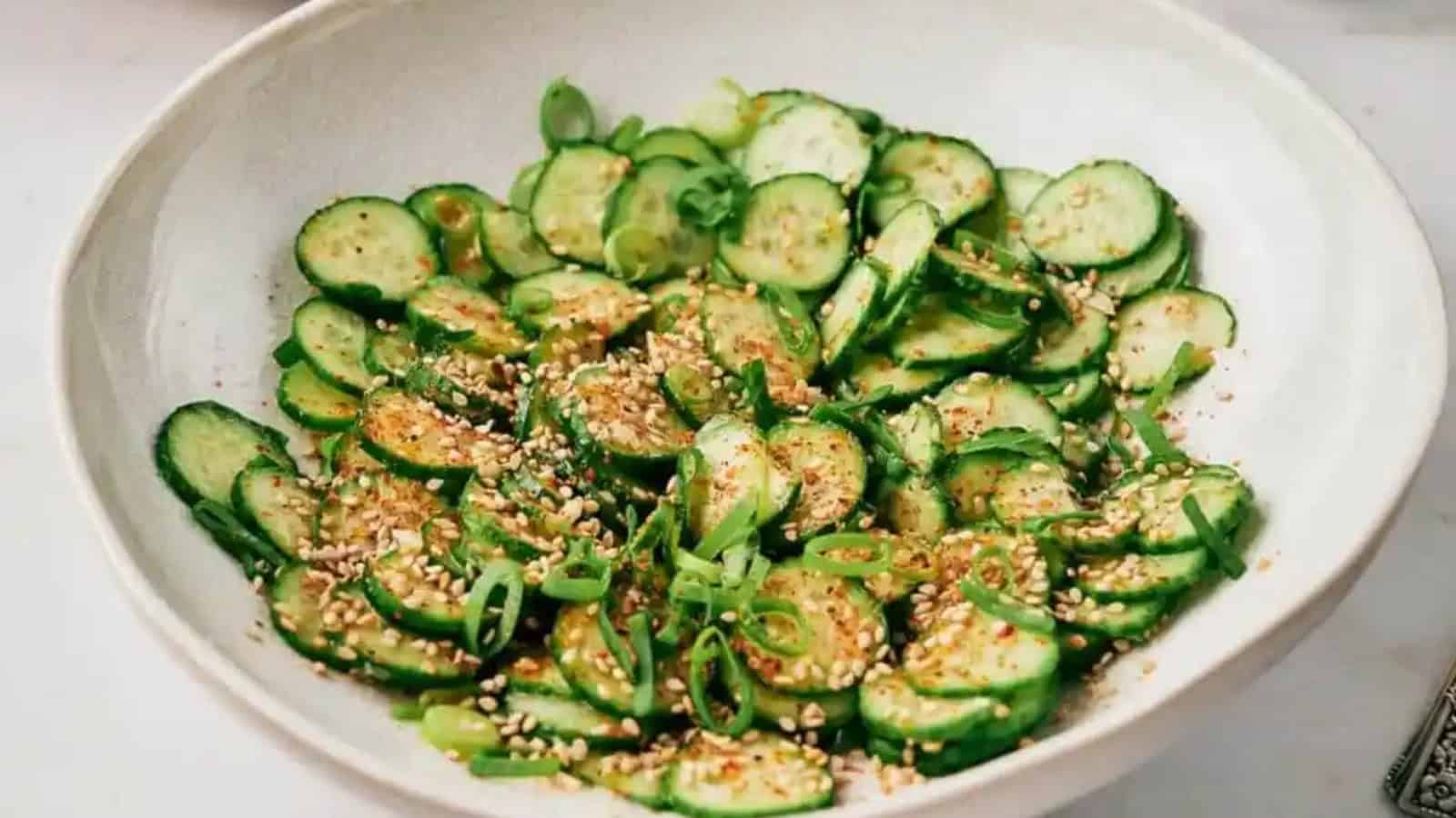 A salad of sliced cucumbers topped with sesame seeds and sliced green onions in a white bowl.