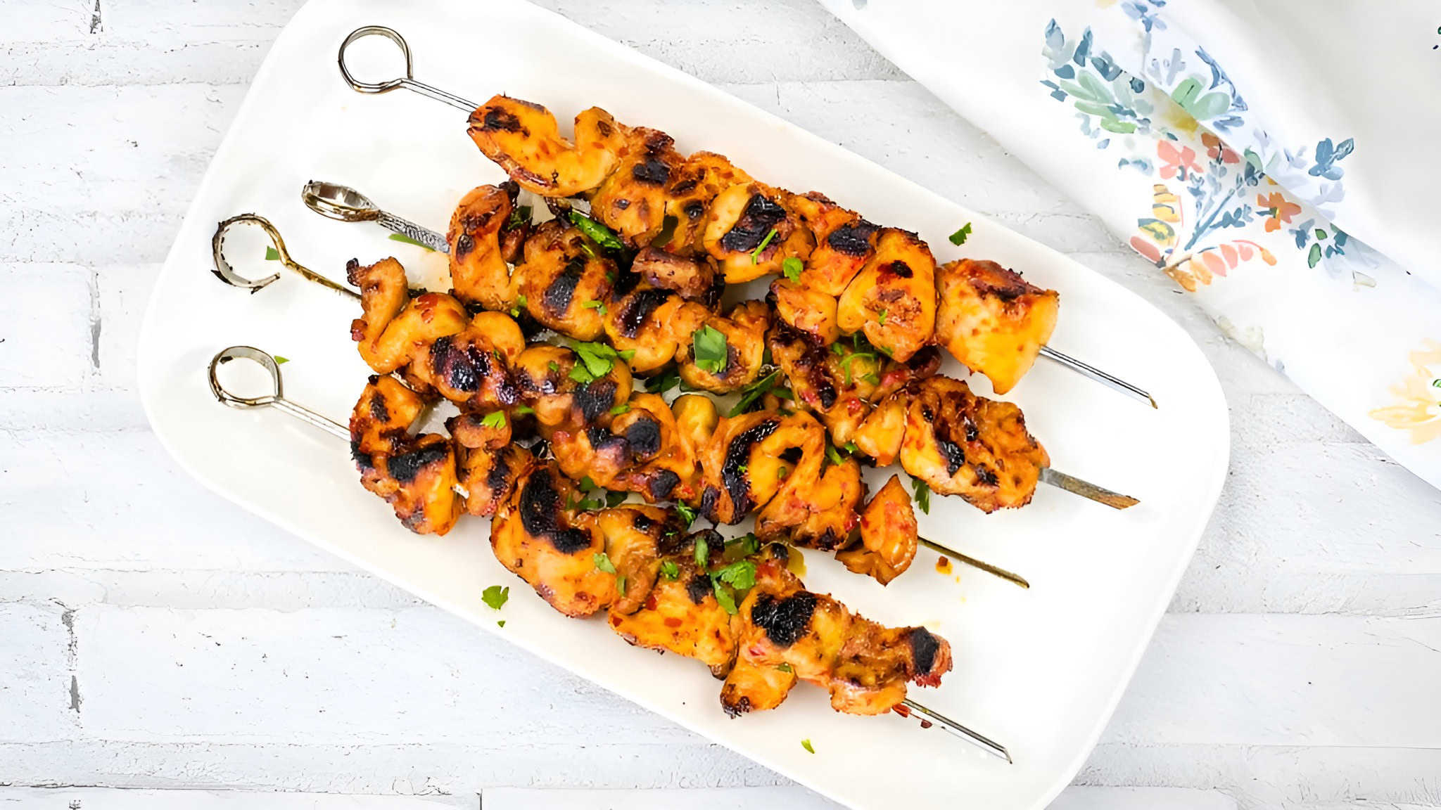 Grilled chicken skewers on a white plate, garnished with herbs. A white floral cloth is partially visible in the background.