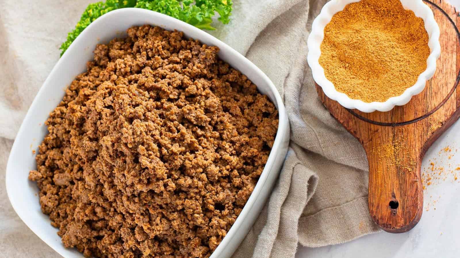 A bowl of seasoned ground beef next to a small dish of taco seasoning on a wooden board. Fresh parsley is seen in the background.