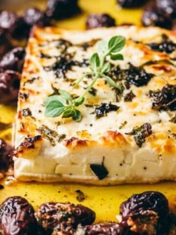 Baked feta cheese with herbs, olives, and a drizzle of olive oil in a baking dish.
