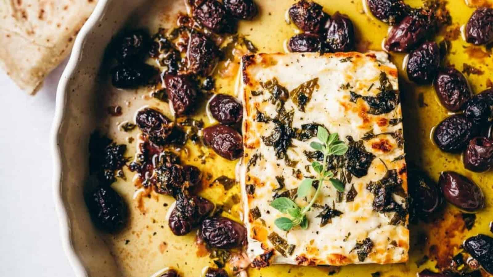 A baked block of cheese garnished with herbs, surrounded by olives in a round dish, with a piece of flatbread on the side.