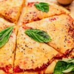 A cheese pizza cut into slices, topped with fresh basil leaves, on a wooden surface with additional basil around it.