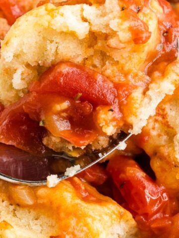 A spoon scooping a serving of tomato cobbler with biscuit topping, featuring chunks of juicy tomatoes and a golden, crumbly biscuit crust.
