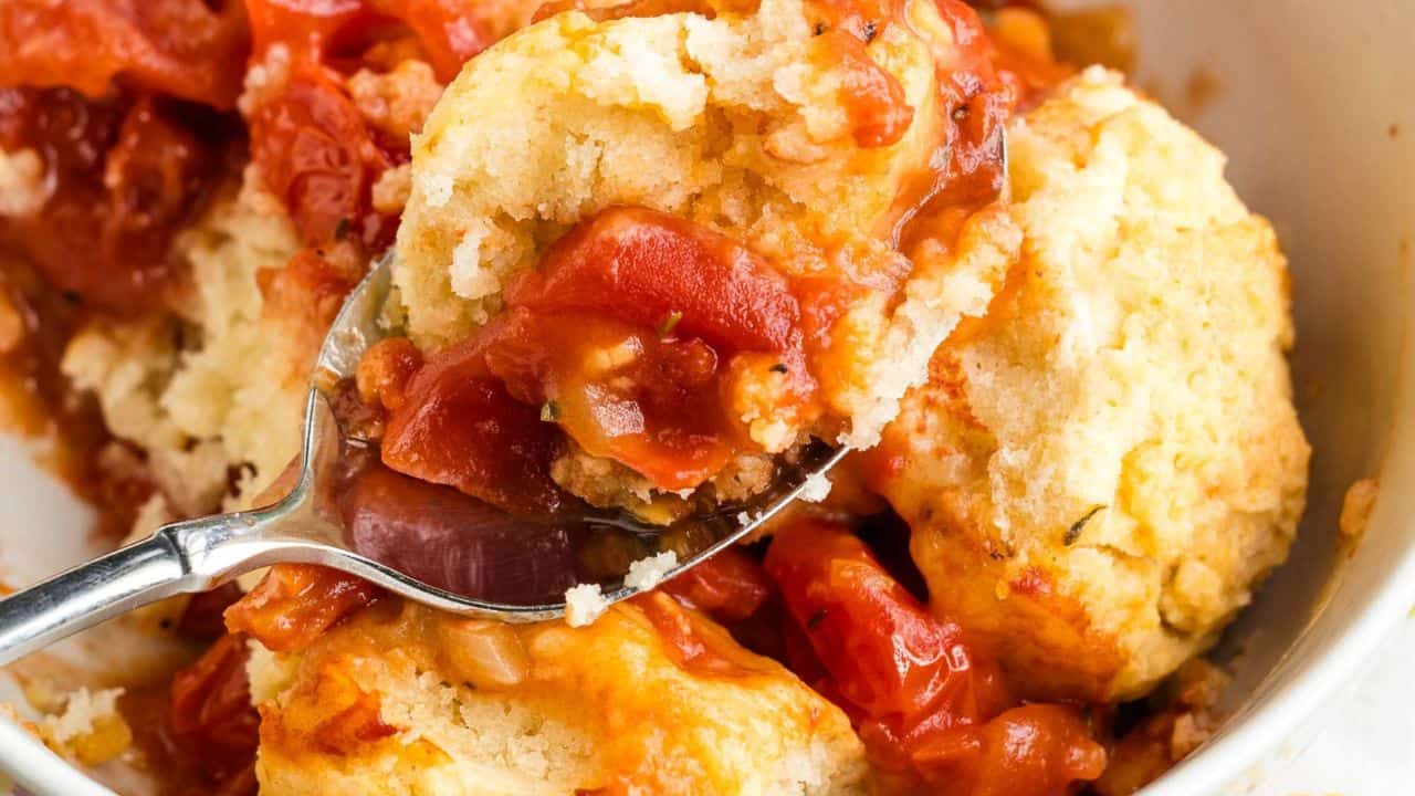 A spoon scooping a serving of tomato cobbler with biscuit topping, featuring chunks of juicy tomatoes and a golden, crumbly biscuit crust.