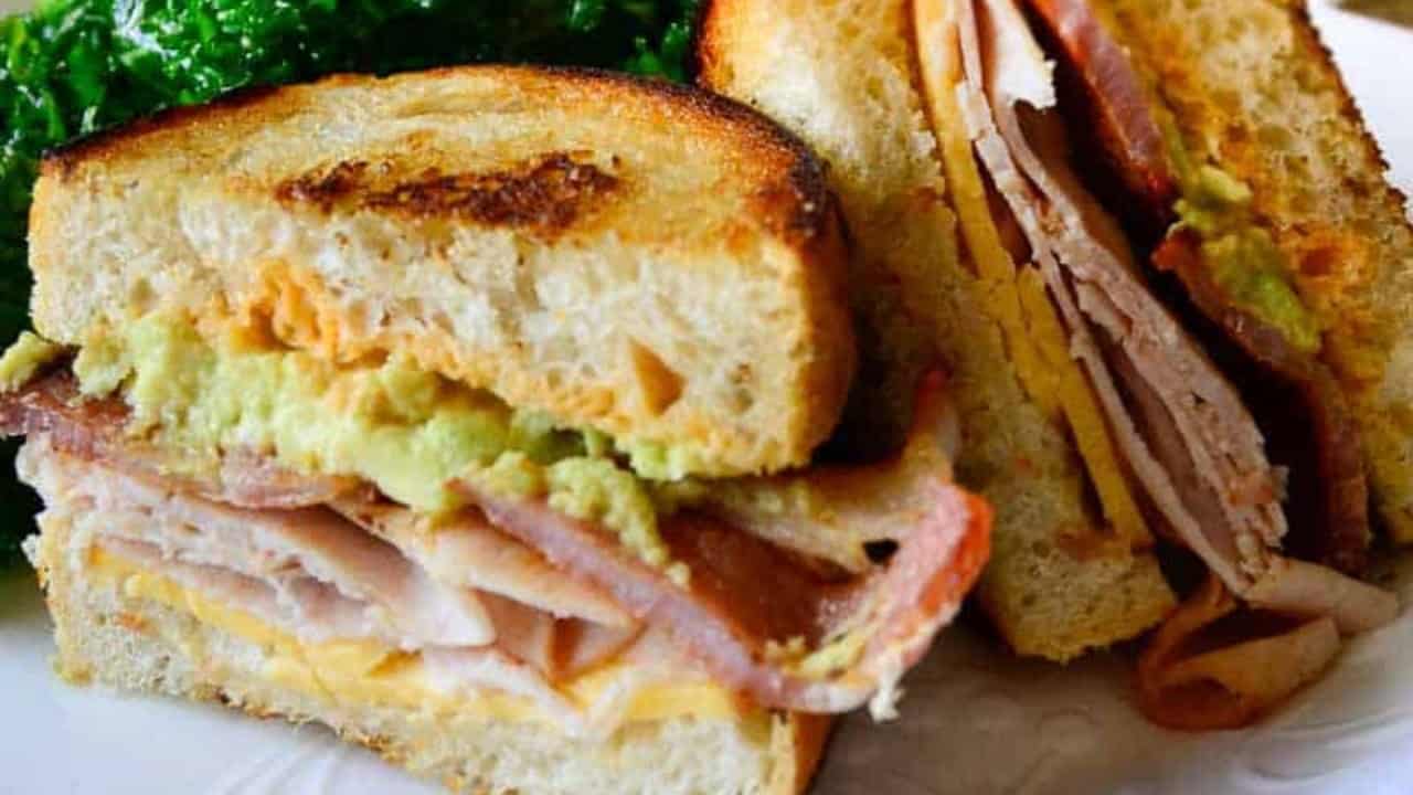 A toasted sandwich with layers of turkey, bacon, cheese, and avocado, served on a plate with a side of leafy greens.