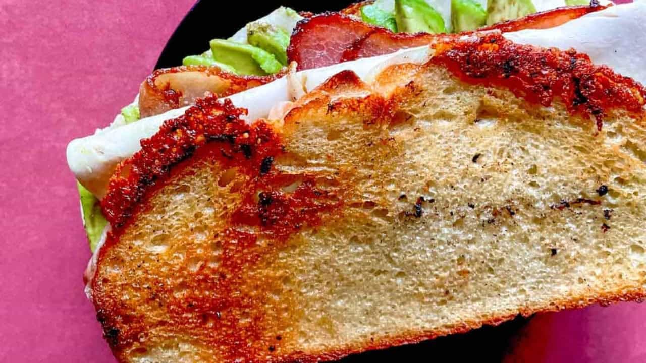 Close-up of a sandwich with toasted bread, turkey slices, avocado, and crispy bacon against a pink background.