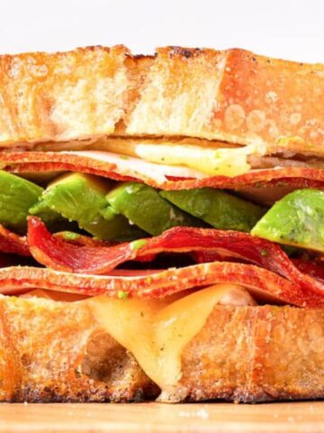 A sandwich with crispy bacon, avocado slices, and melted cheese between toasted bread slices.