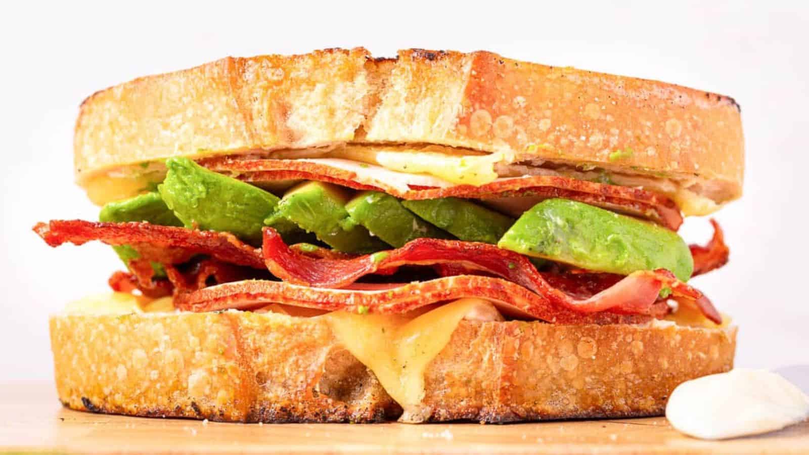 A sandwich with crispy bacon, avocado slices, and melted cheese between toasted bread slices.