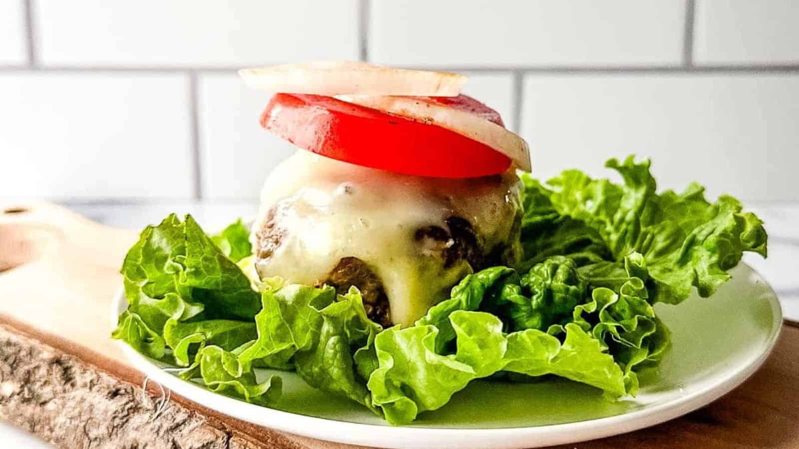 A cheeseburger patty with cheese, tomato, and onion slices sits on lettuce on a white plate.