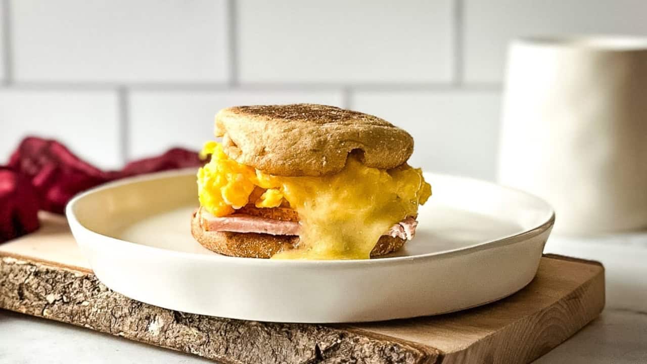 Breakfast sandwich with scrambled eggs, cheese, and ham on an English muffin, served on a white plate placed on a wooden board.