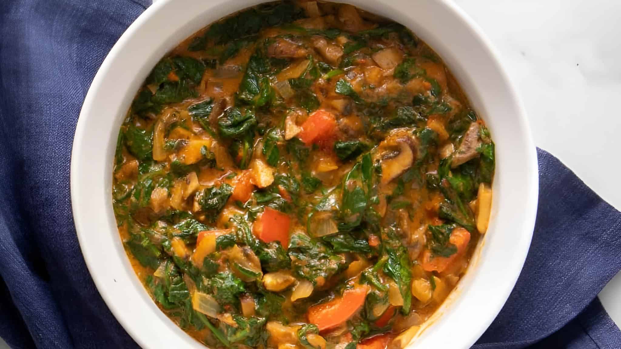 A bowl of colorful vegetable stew with spinach, tomatoes, carrots, and other vegetables on a dark blue cloth.