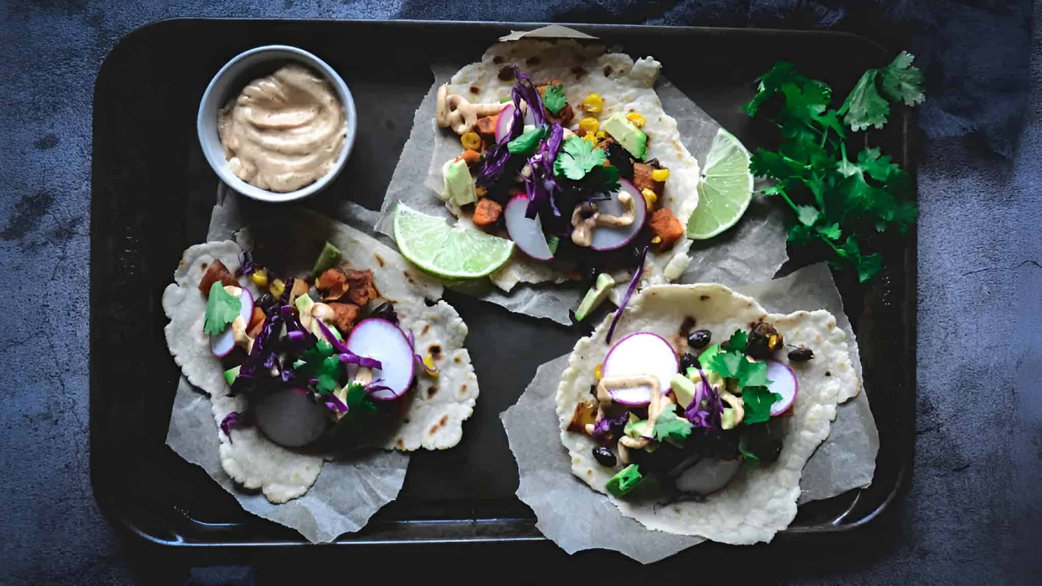 Three corn tortillas topped with assorted vegetables, radishes, and cilantro, served with a lime wedge and a small bowl of sauce on a dark tray.