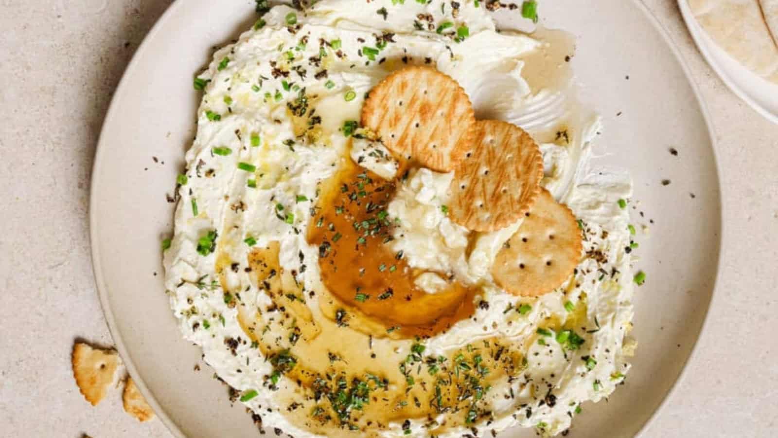 A plate of creamy dip topped with herbs, olive oil, and three round crackers on a neutral-toned surface.