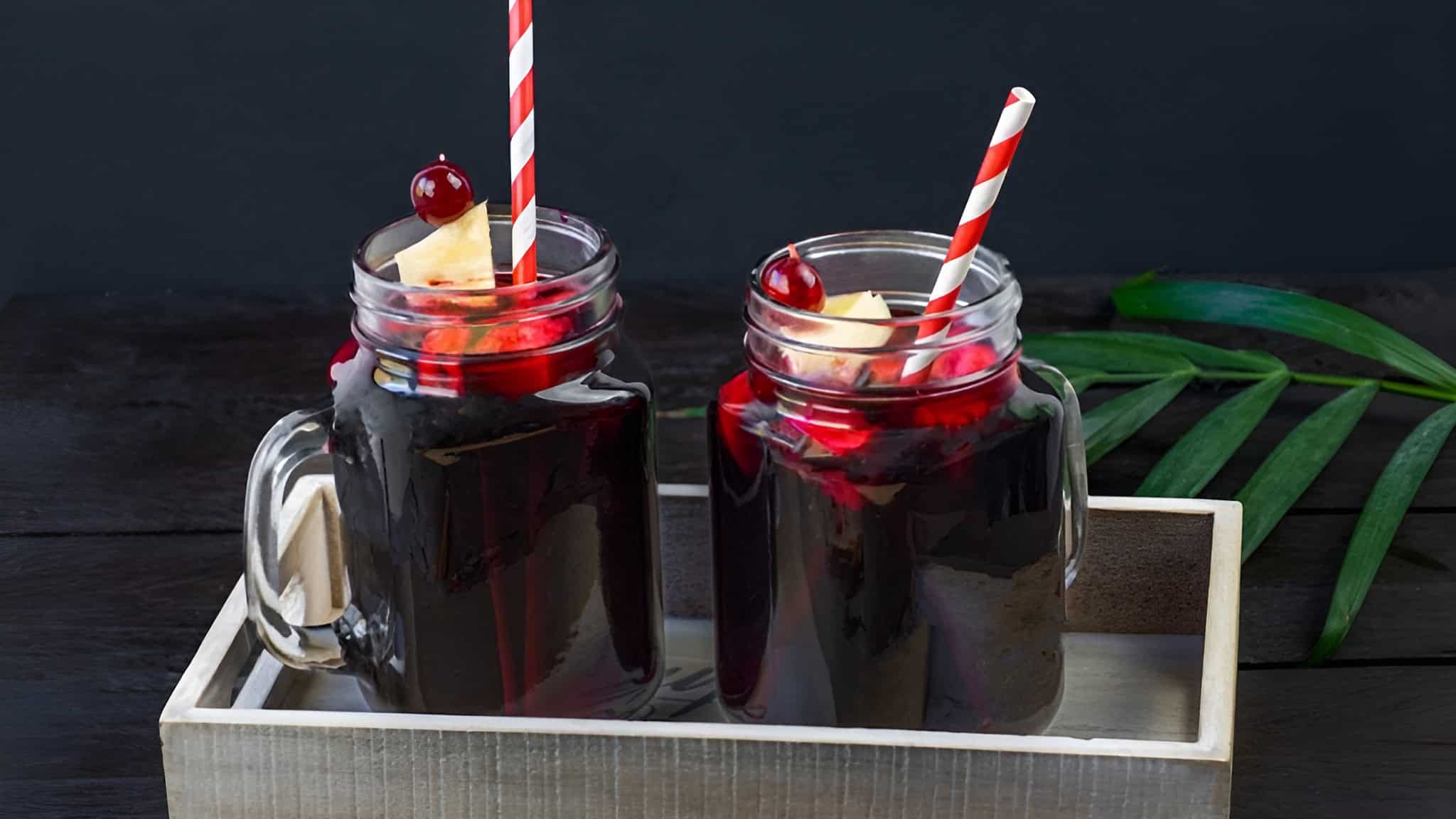 Two mason jars filled with dark red liquid, topped with cherries and pineapple slices, each with a red-striped straw, placed on a wooden tray beside a green leaf.
