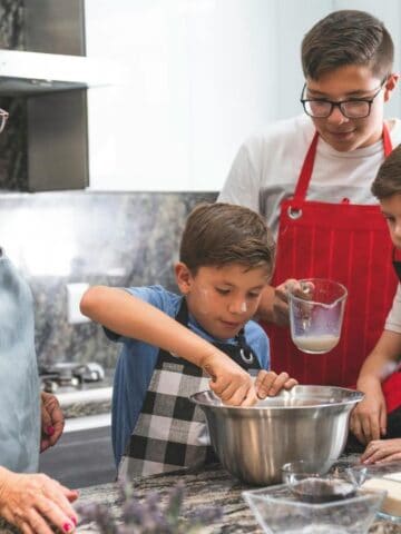 A woman and three boys wearing aprons are baking together in a kitchen, mixing ingredients in a metal bowl on a countertop with eggs and other baking items nearby.