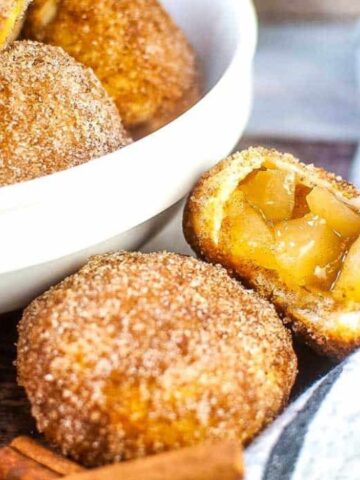 A bowl of cinnamon sugar-coated pastries, with one pastry cut open to show an apple filling. Striped cloth and butter in the background.
