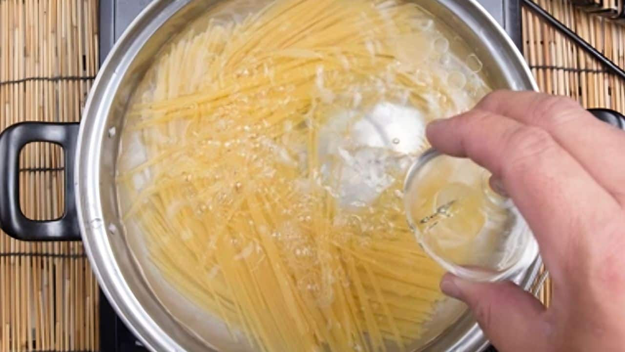 A hand pours liquid from a small glass into a pot of boiling pasta on a stovetop.
