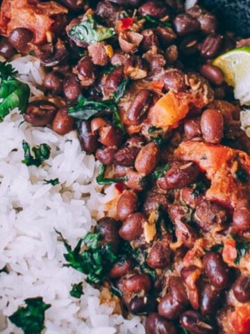 A black bowl with cooked white rice, black beans with vegetables, lime wedges, and a spoon.