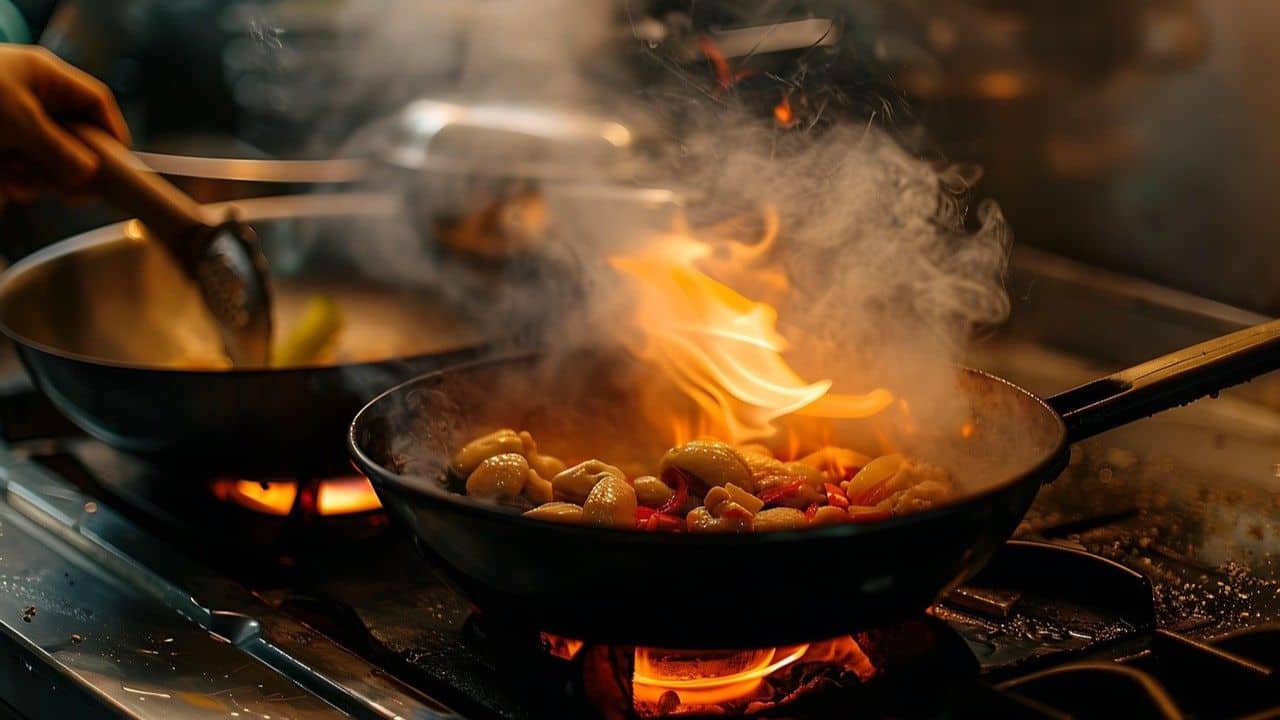 A person cooking vegetables and beans in a pan over a flame on a stovetop, with steam and a small flare-up visible.