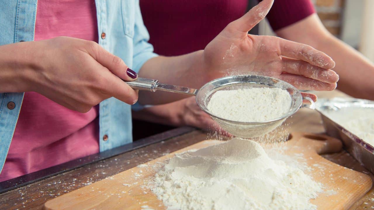 Two people sifting flour onto a wooden board using a metal sieve.