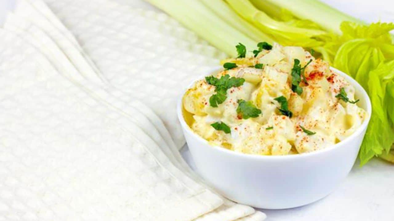 A bowl of creamy potato salad garnished with chopped parsley and paprika, next to celery stalks and a folded white cloth.