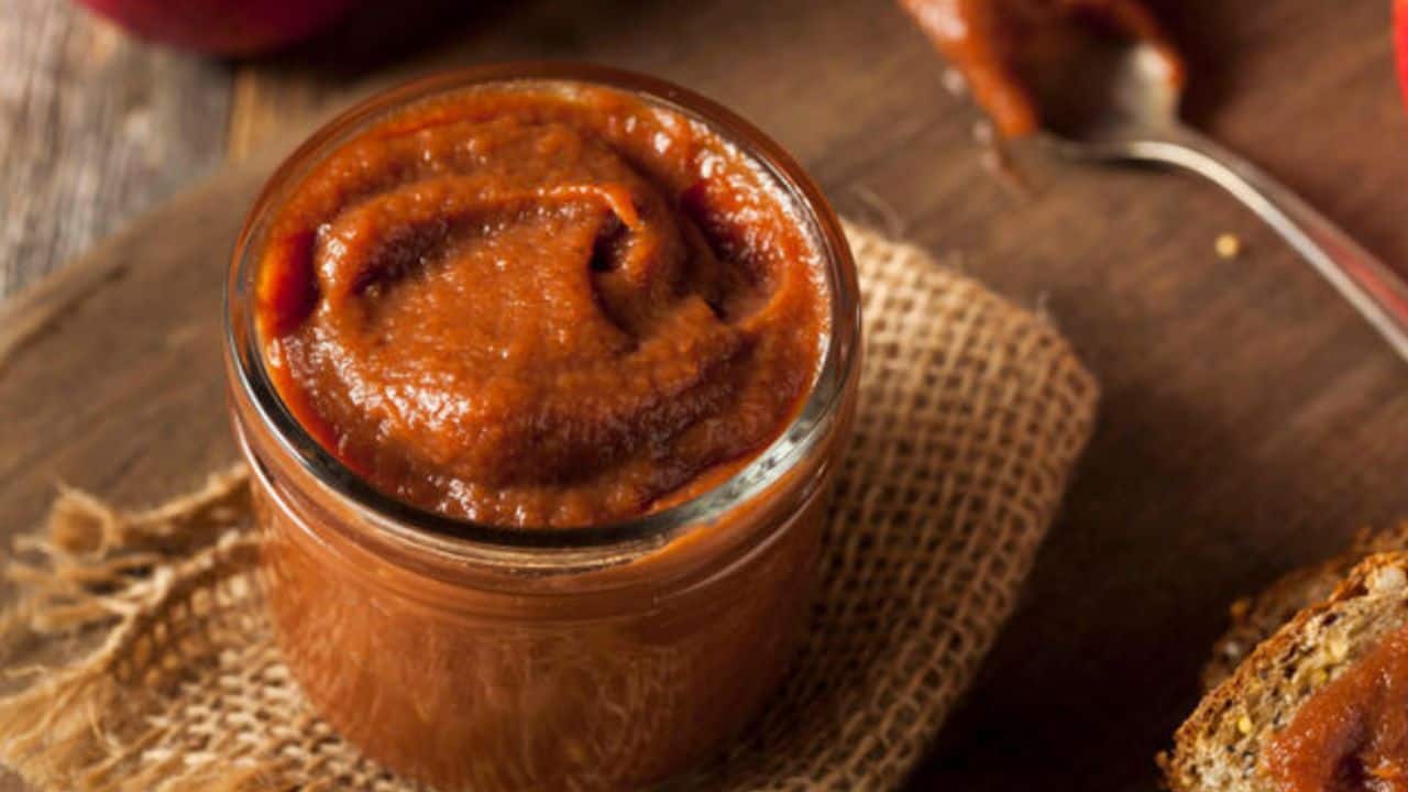 A jar of apple butter on a burlap cloth with a spoon in the background.