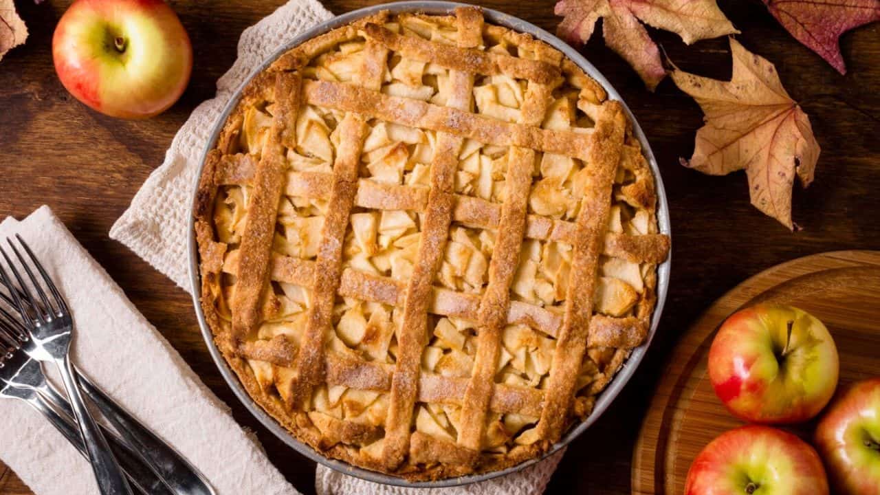 A freshly baked apple pie with a lattice crust sits on a wooden table next to apples, autumn leaves, and utensils wrapped in a napkin.