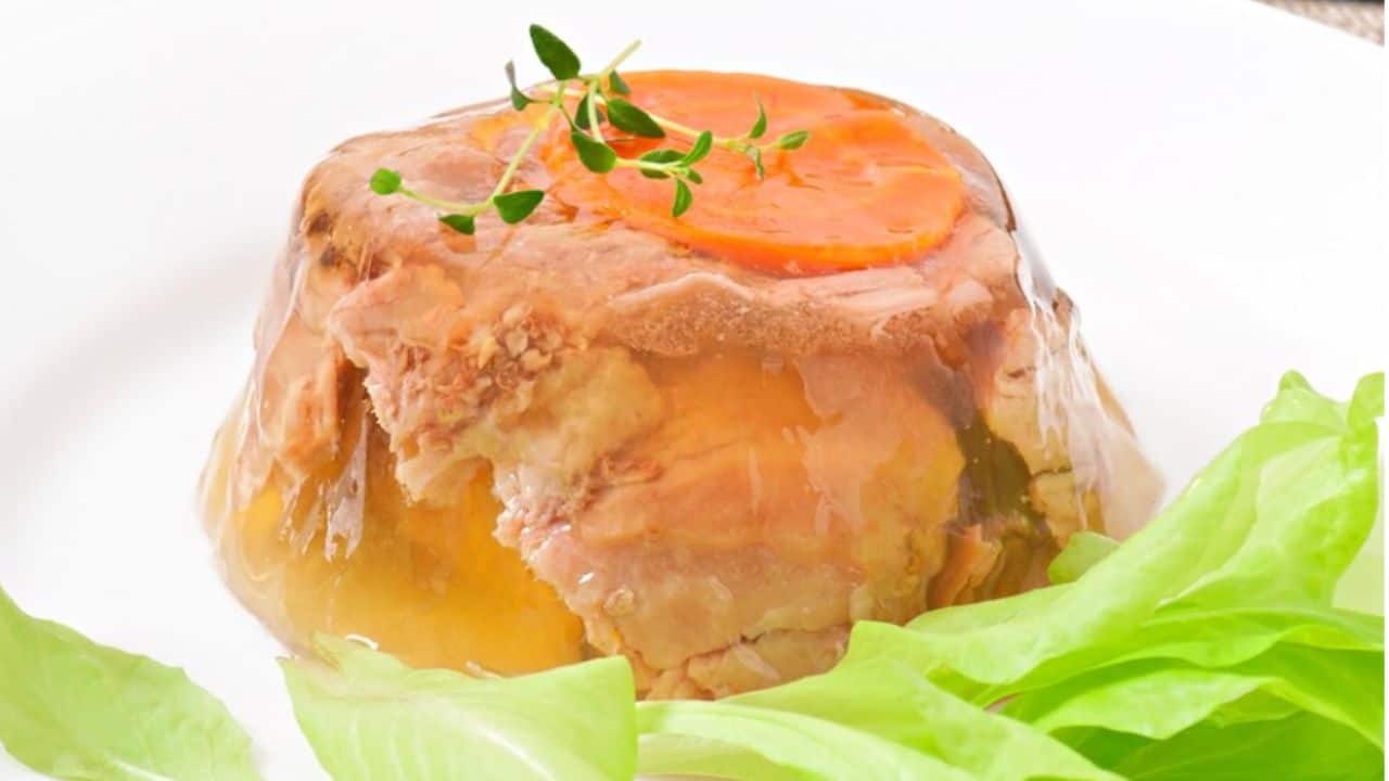 A meat aspic with a slice of carrot on top, garnished with a sprig of herbs, served on a white plate next to fresh lettuce leaves.