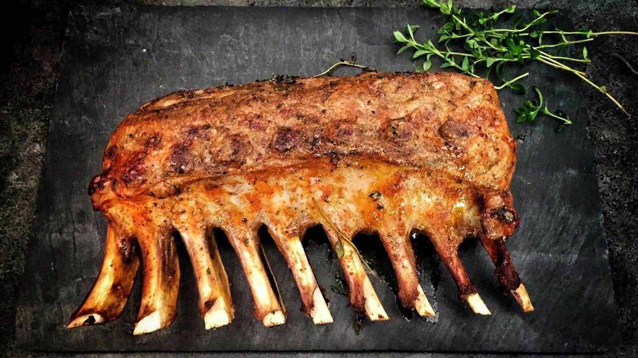 A cooked rack of ribs is placed on a dark slate surface, garnished with fresh sprigs of herbs.