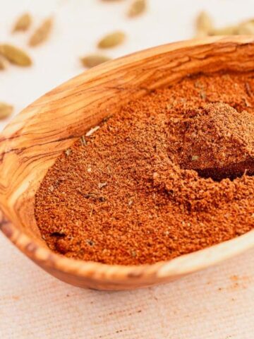 A wooden bowl filled with ground spices and a wooden spoon; scattered whole spices are in the background.