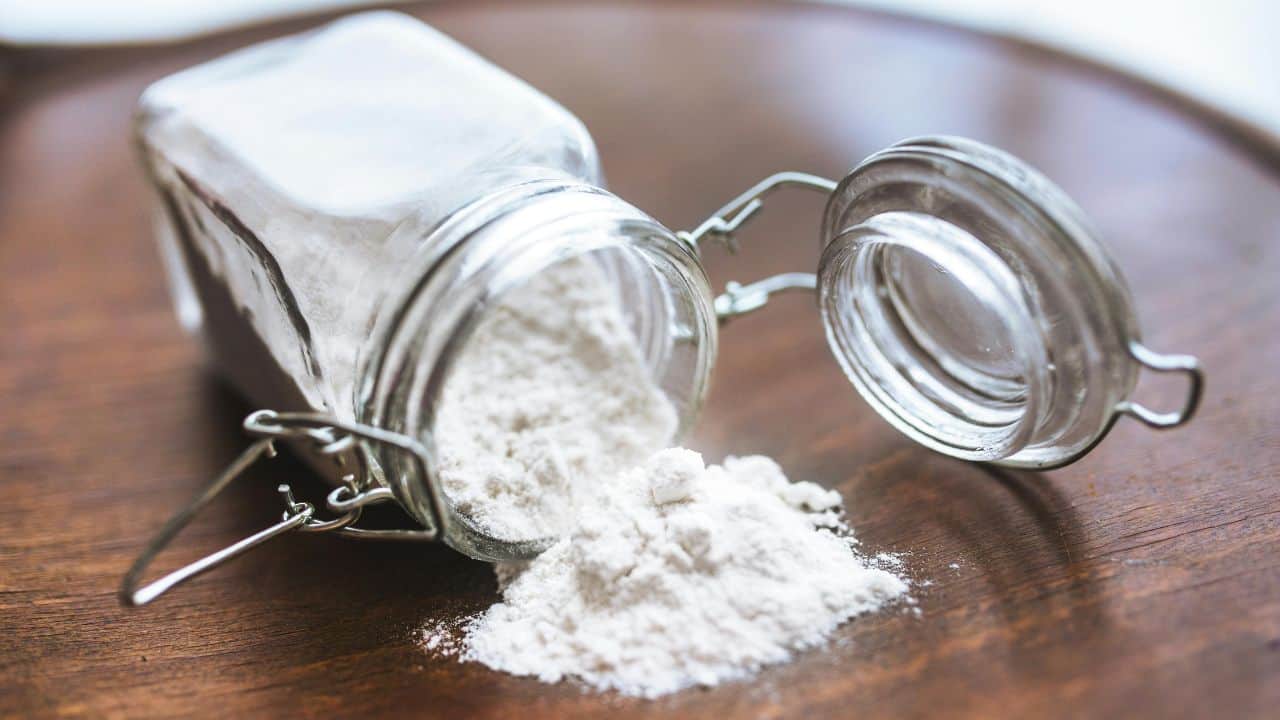 Open glass jar with white flour spilled onto a wooden surface.