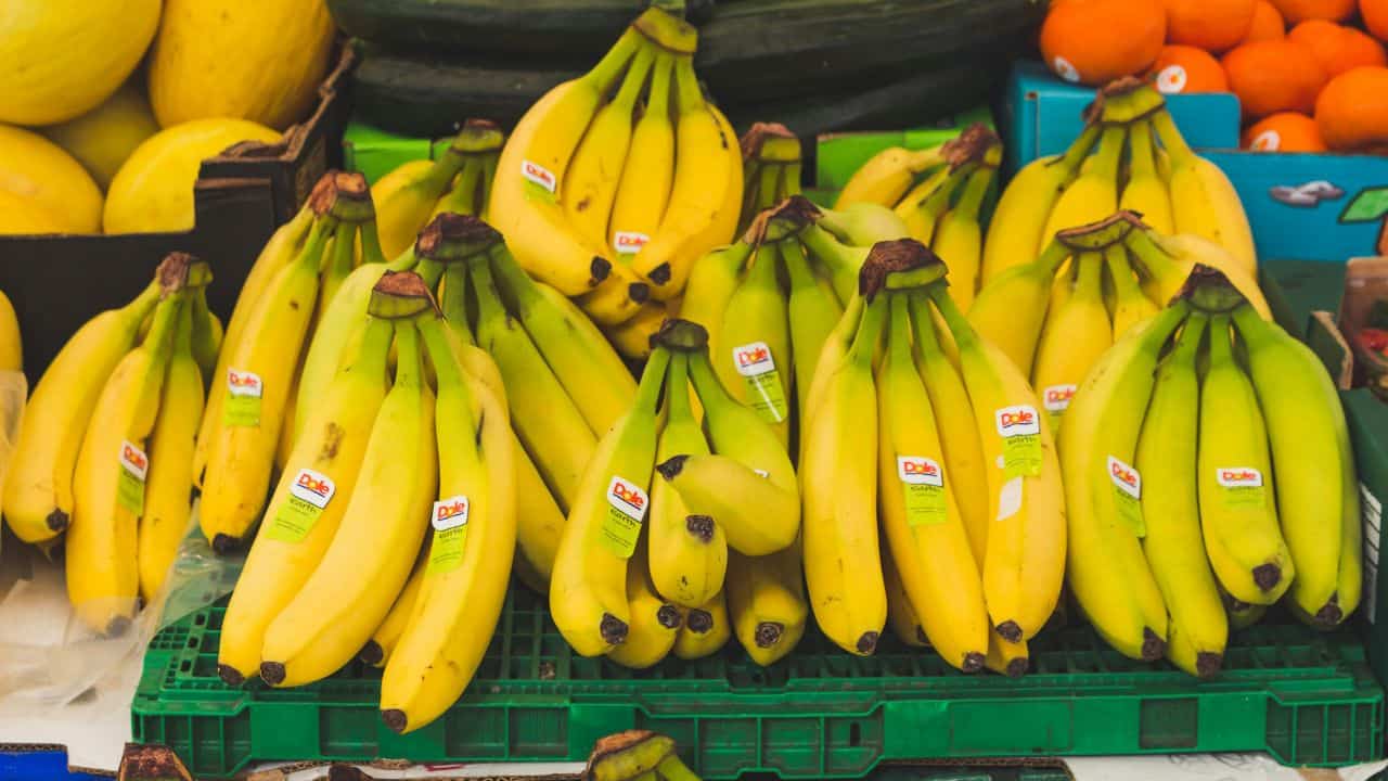 Bunches of ripe bananas with stickers are displayed on a green plastic crate at a market stall, surrounded by other fruits.