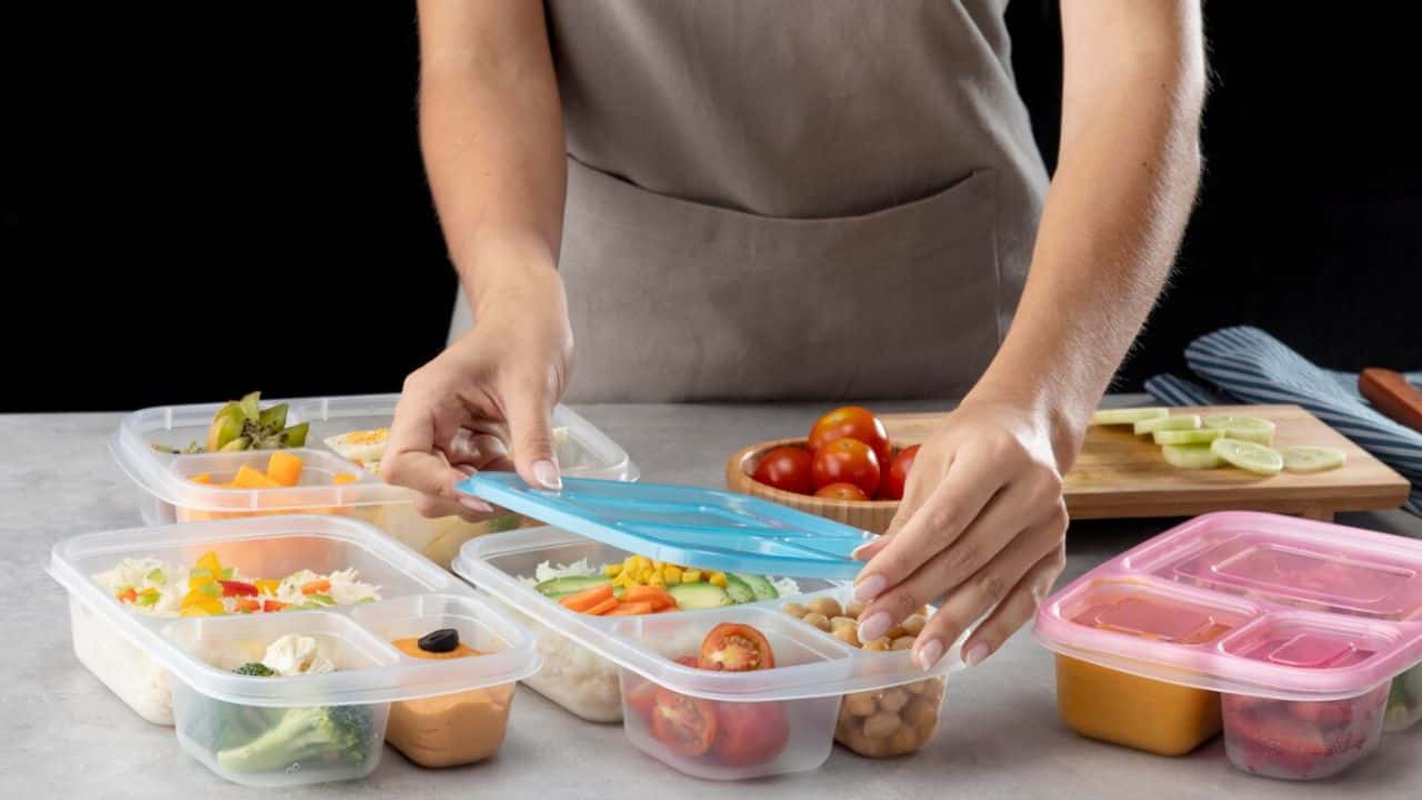 Person organizing meal prep containers with various salads and vegetables on a table, alongside a cutting board with tomatoes and cucumbers.