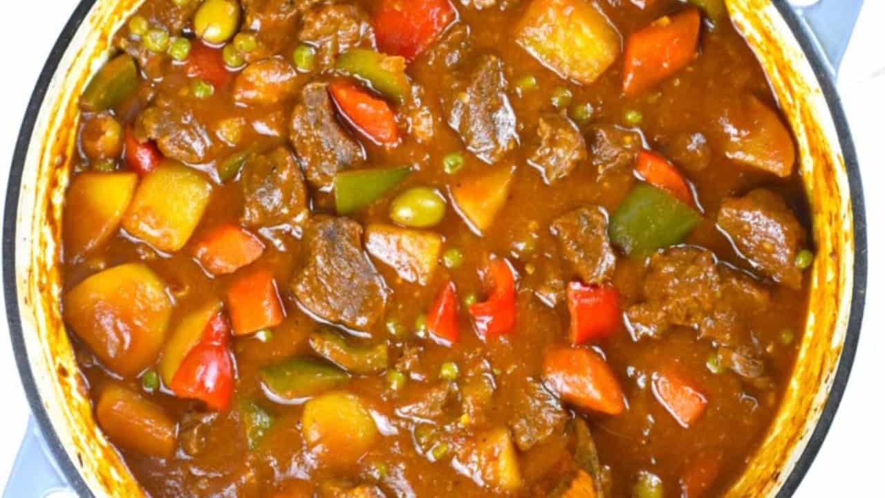 A pot of beef stew with chunks of meat, potatoes, carrots, peas, and bell peppers in a rich, brown sauce.