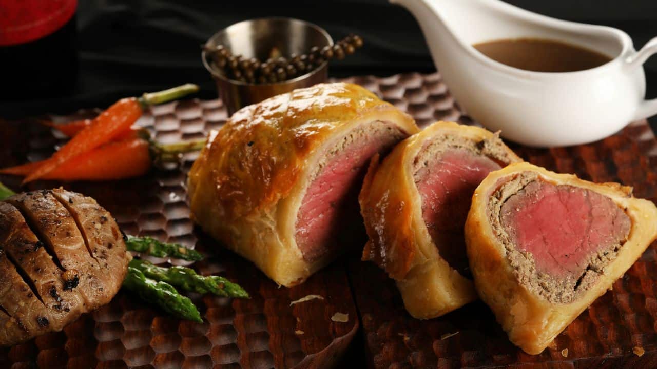 Sliced beef Wellington with asparagus, carrots, a meatball, and sauces on a wooden board.