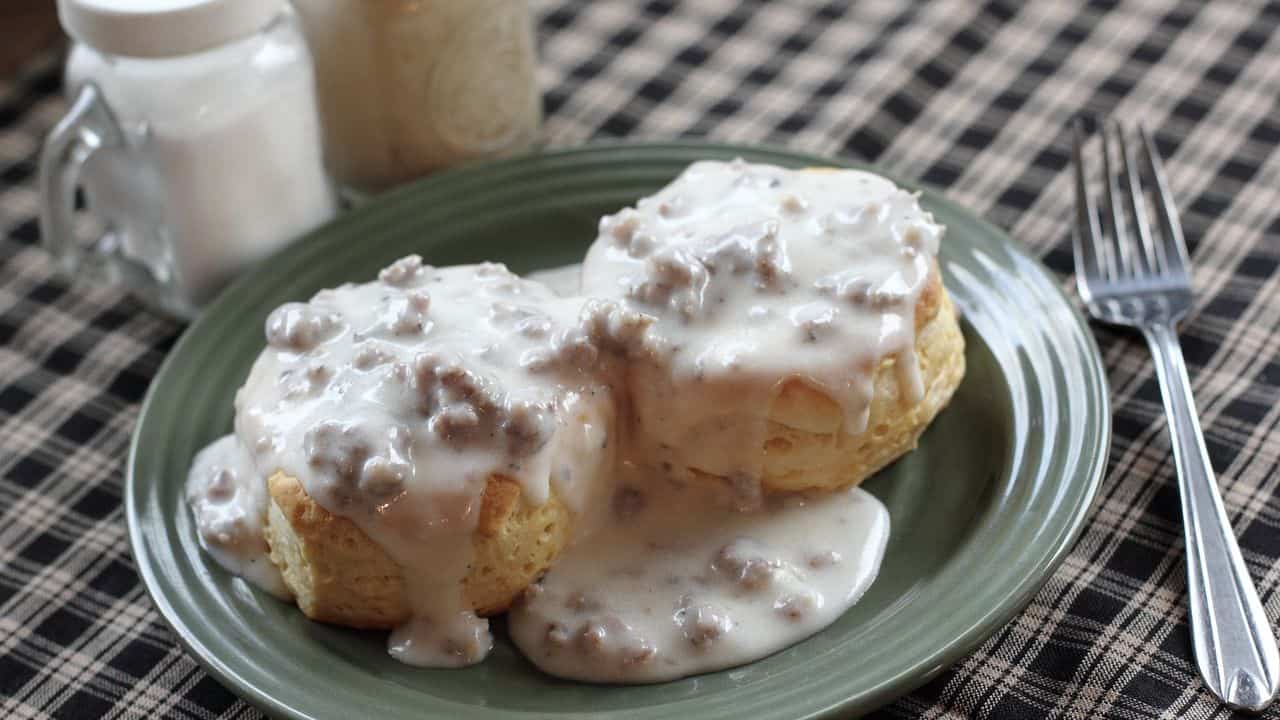 Two biscuits topped with sausage gravy on a green plate, with salt, pepper, and a fork on a checkered tablecloth background.