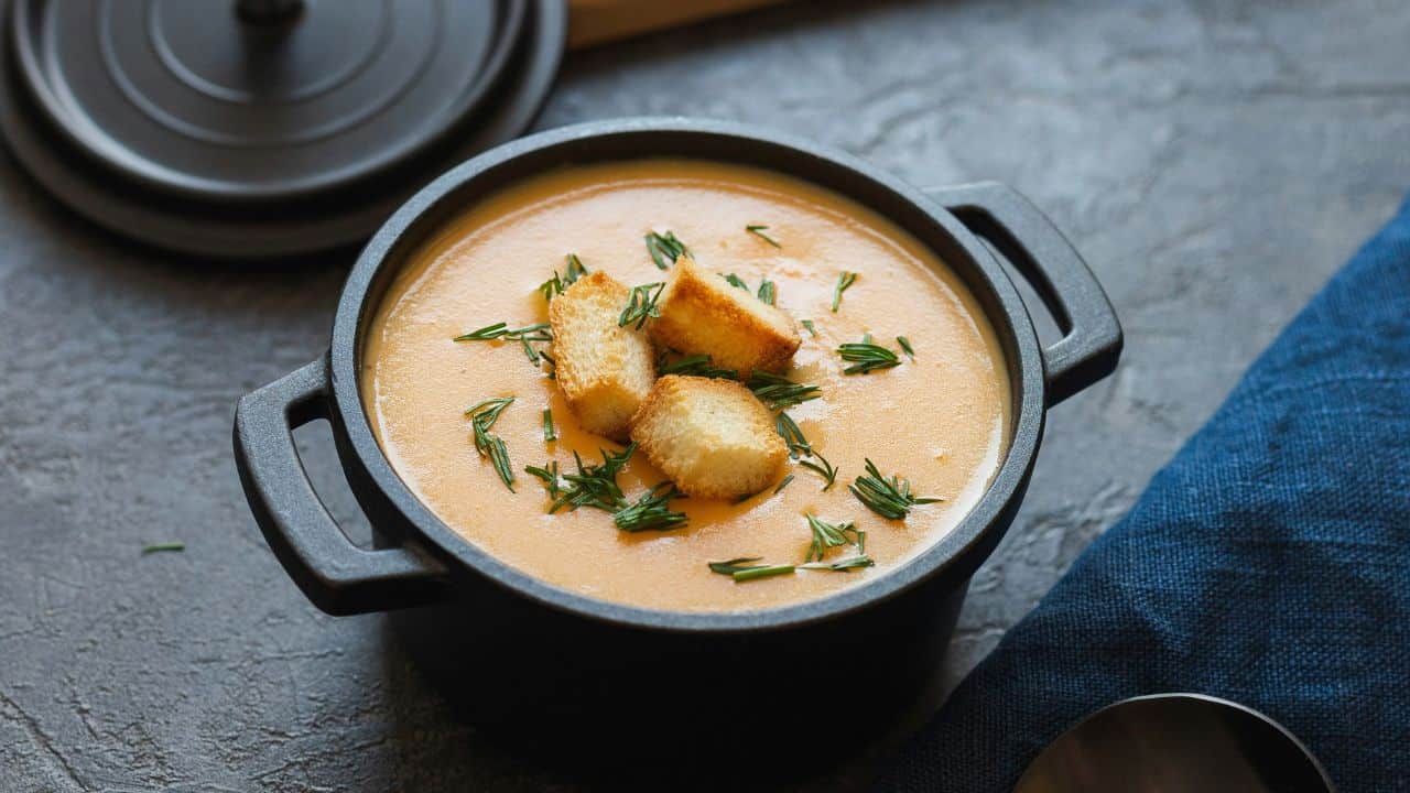 A bowl of creamy soup topped with croutons and herbs, placed on a gray surface with a lid and dark blue cloth nearby.