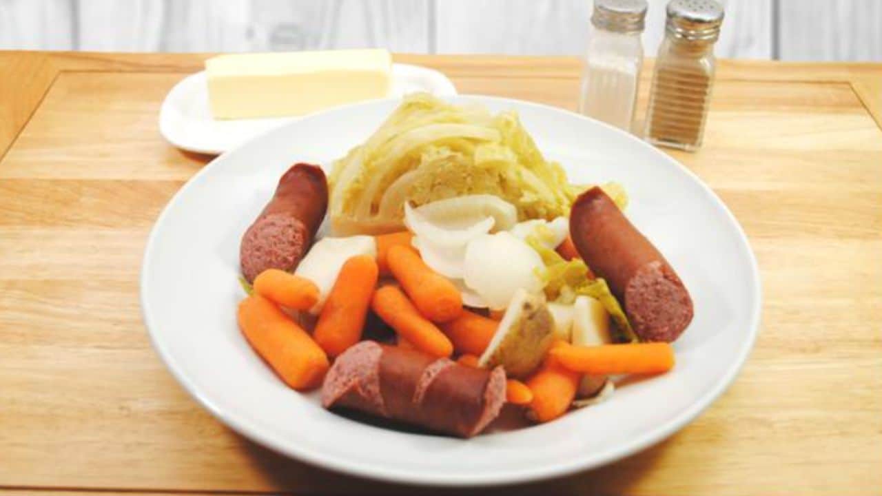 A plate with sliced sausage, carrots, cabbage, and potatoes. Butter and salt and pepper shakers are in the background.