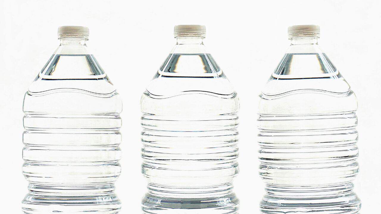 Three large, transparent plastic water bottles with white caps are lined up in a row against a plain background.